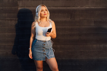 Sexy young woman with long blonde hair wears fashionable clothes and listens to current music in the street on a black stone background.