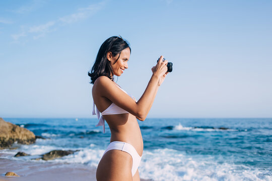 Side view of cheerful Latin woman in bikini swimsuit enjoying photo hobby while testing vintage camera and making images of ocean horizon, happy female swimmer in white bikini using retro technology