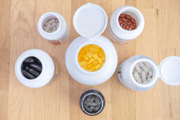 Open jars with yellow omega 3 capsules, red lutein, black ubiquinol, with white vitamins capsules, jars with sports and health supplements on a wooden surface top view