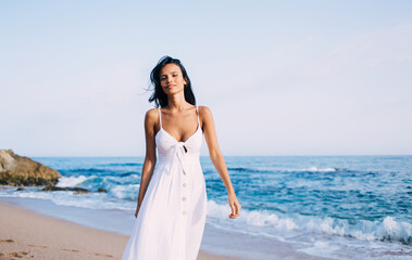 Fototapeta na wymiar Portrait of Latin woman dressed in comfortable white sundress walking at coastline beach during pastime near Caribbean sea, charming female model looking at camera during promenade on vacations