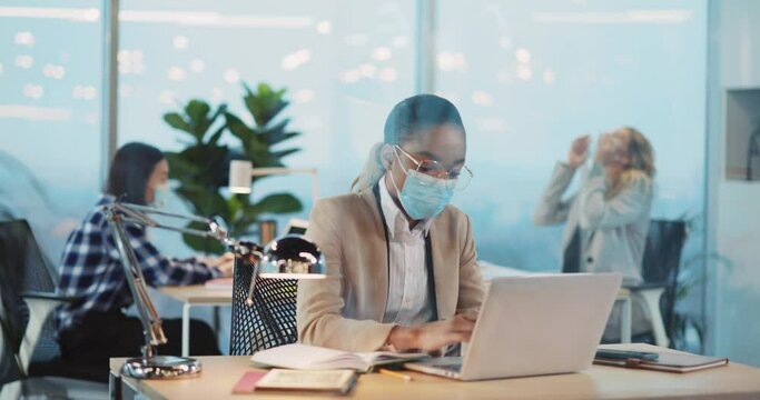 African young woman in mask working in central office during quarantine. Young sick blonde executive sneezing allergy calming up colleagues.