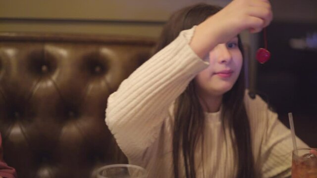 Happy Young Girl at Restaurant Booth Sips on Iced Tea and Eats Maraschino Chery
