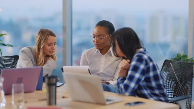 Pretty sociable multi-cultural young women discussing computer project on laptop negotiating business plans coworking in corporate modern office.