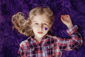 International world epilepsy illness awareness day. Cute pretty blonde Caucasian girl with small violet purple paper heart on cheek lying on purple fluffy rug carpet at home. View from above. - 415020963