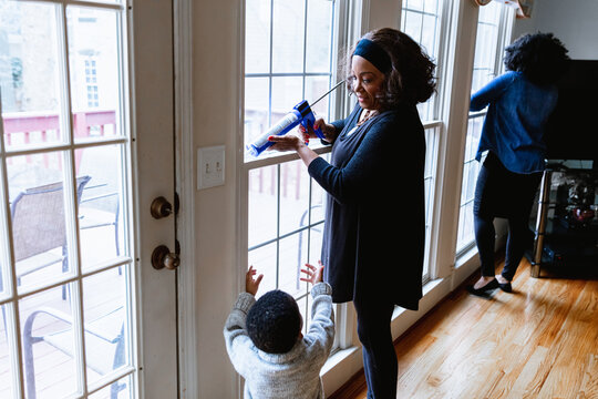 Black grandmother woman weather sealing, caulking windows for winter with toddler grandson