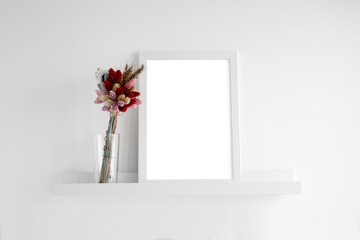 White frame on a white shelf on a white wall. On the shelf next to the composition of dried flowers. Abstract minimalistic background