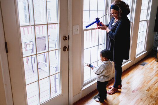 Black grandmother woman weather sealing, caulking windows for winter with toddler grandson