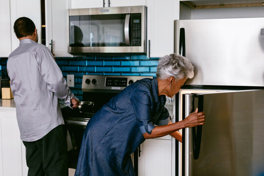 Wife getting drink from refrigerator while husband prepares to cook in kitchen, senior couple
