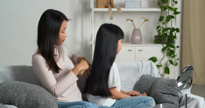 Happy loving asian family. Korean mother wears casual clothing combing her daughter's hair uses comb brush to take care of her brunette daughter sitting on couch in living room. Motherhood family love