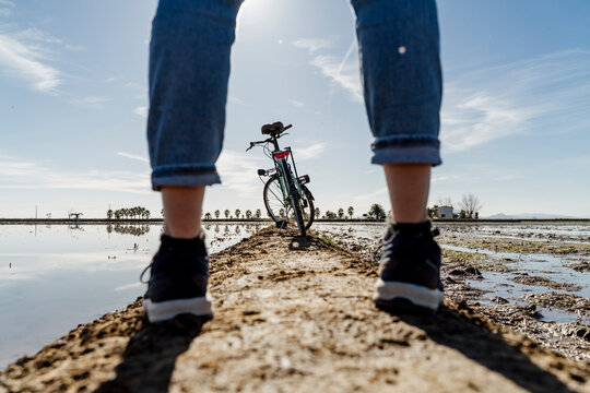 Bicycle seen through legs of woman standing on footpath at Ebro's Delta during sunny day, Spain
