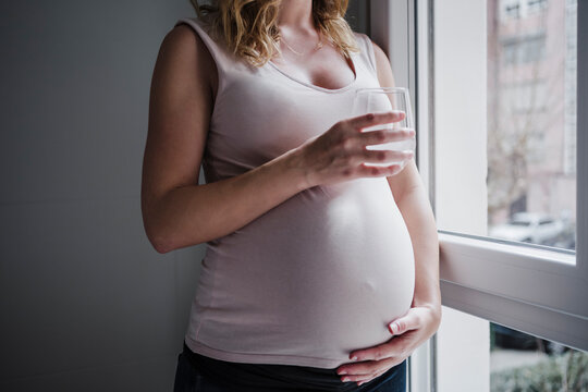 Pregnant woman holding drinking glass while standing by window at home