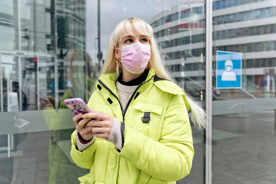 Young woman wearing face mask using smart phone while standing against glass wall