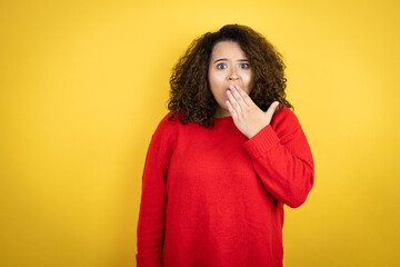 Young african american woman wearing red sweater over yellow background covering mouth with hand,...