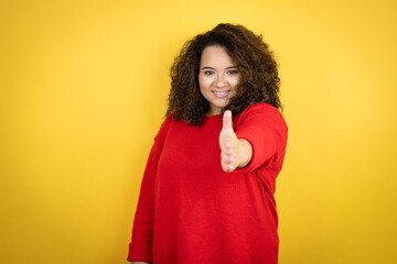 Young african american woman wearing red sweater over yellow background smiling friendly offering handshake as greeting and welcoming