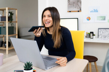 Happy female student with laptop talking on speaker phone while sitting on chair at home