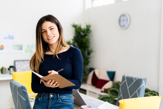 Smiling female student holding notebook and pen while standing in living room