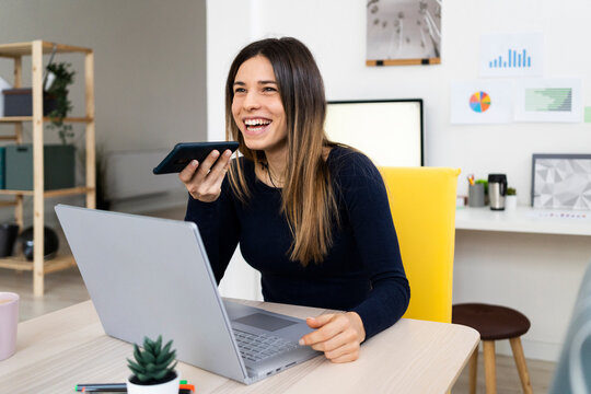 Happy female student with laptop talking on speaker phone while sitting on chair at home