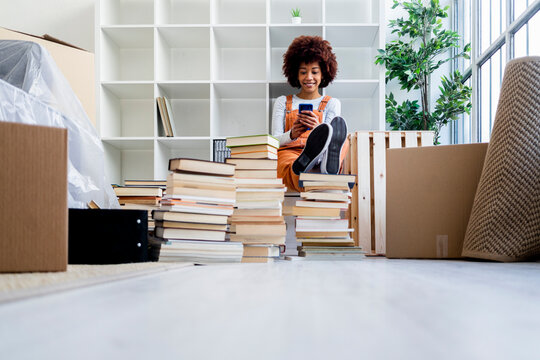 Afro woman using smart phone while siting against bookshelf in new home