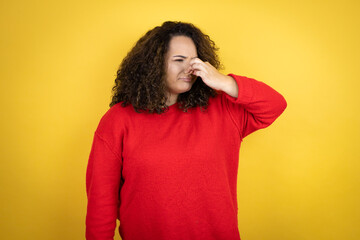 Young african american woman wearing red sweater over yellow background smelling something stinky and disgusting, intolerable smell, holding breath with fingers on nose