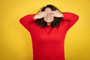 Young african american woman wearing red sweater over yellow background covering eyes with hands smiling cheerful and funny. Blind concept.