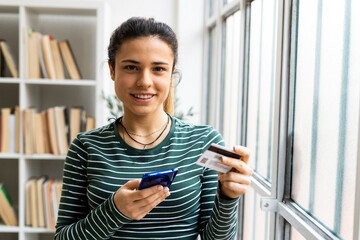 Smiling young woman with smart phone and credit card standing by window at work place