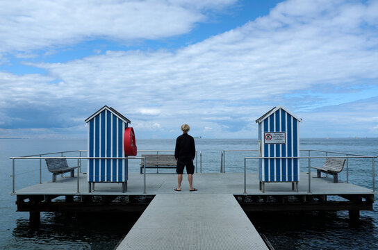 Germany, Schleswig Holstein, Stein, Rear view of man standing on jetty with blue and white beach cabins on Baltic Sea under cloudy sky