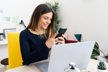 Smiling freelancer worker using mobile phone while sitting by laptop in living room