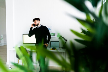 Businessman talking on smart phone in office, green plant leaves in foreground