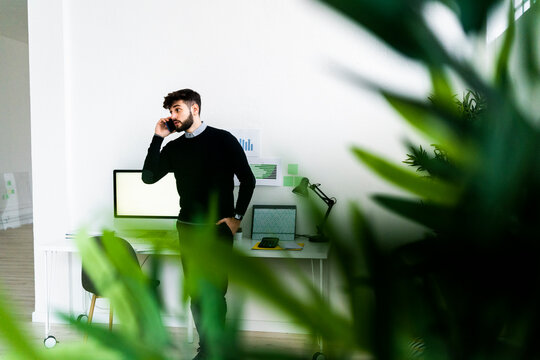 Businessman Talking On Smart Phone In Office, Green Plant Leaves In Foreground