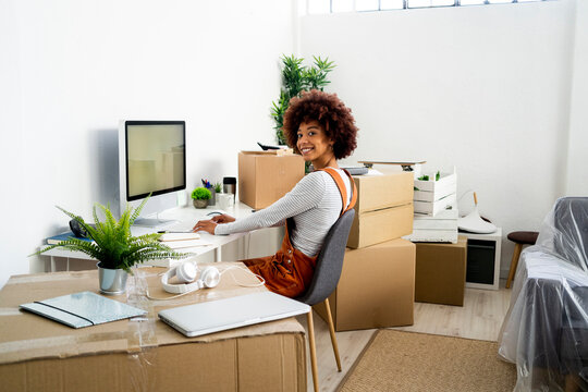 Smiling woman sitting on chair by computer while moving in new apartment