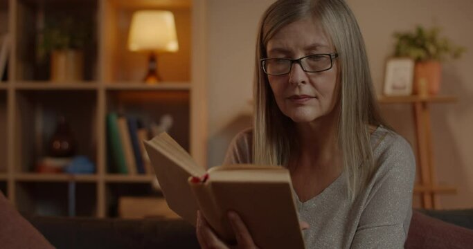 Close up of senior woman turning page while reading and spending free time. Atractive female person in glasses enjoying book while sitting on sofa at home. Concept of retirement, leisure.