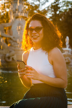 Smiling woman wearing sunglasses using smart phone while sitting against fountain during sunset