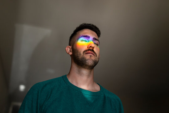 Rainbow light hitting face of young man standing indoors with closed eyes
