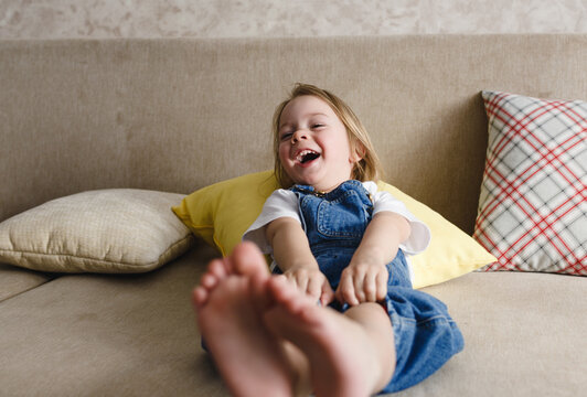 a little girl in a blue jumpsuit lies at home on the sofa among the colorful pillows and smiles cheerfully