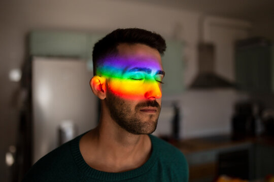 Rainbow light hitting face of young man standing indoors with closed eyes