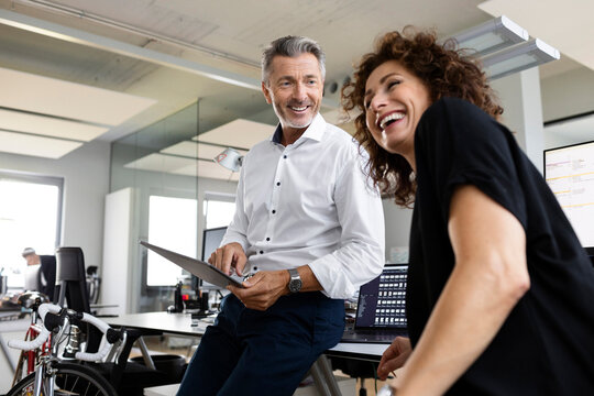 Smiling businessman with digital tablet looking at cheerful colleague while standing at open plan office