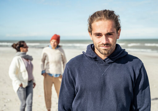 Portrait of young man wearing hooded shirt standing on beach with two young woman walking behind in background