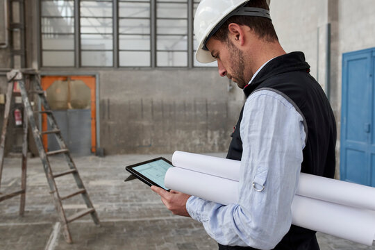 Male architect with blueprints using digital tablet while standing in building