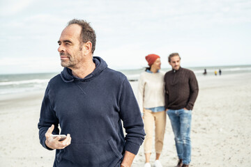 Portrait of man standing on sandy beach with smart phone in hand with young couple in background