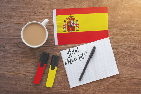 spain national flag, notebook with the inscription hello, how are you in spanish, two markers, cup of coffee on brown wooden desktop, study concept 