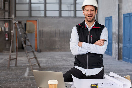 Male architect with arms crossed laughing while standing at table in building