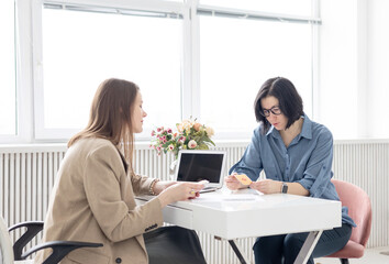 two young women conduct a coaching session in home office in light interior with laptop and flowers on tablework with metaphorical cards and psychological counseling