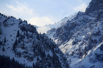Sharp rocks in the high-mountain gorge of the Medeo tract under snow in the early morning in winter, trees on the rocks, the sun illuminates the rocks, the sky with clouds