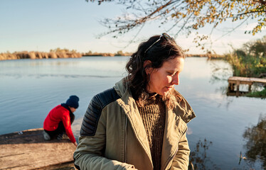 A mother in the foreground walks towards the camera and her son in a hat and red shirt is playing with the lake water on the pier