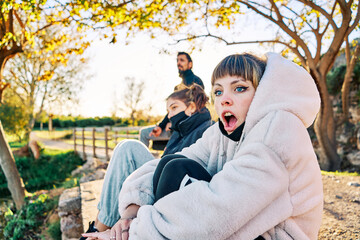 Fototapeta premium Young girl with blue eyes in the foreground with surprised expression enjoying the sunset in the park with her friends