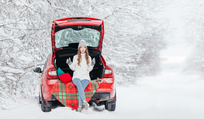 A woman with hot coffee in her hands sits in a red car on a snowy winter day in the forest.
