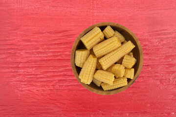 Top view of a wood bowl filled with canned organic baby corn offset on a red painted background.