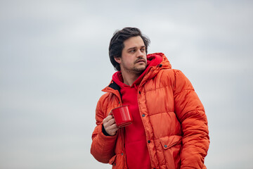 Hipster guy with red mug drink a coffee in outdoor with clouds on background