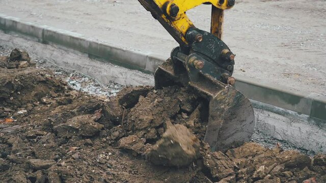 Excavator arm scoops up a bucket full of soil and rocks during roadworks in the countryside. Close up view of a big yellow excavator digging a deep trench near construction site
