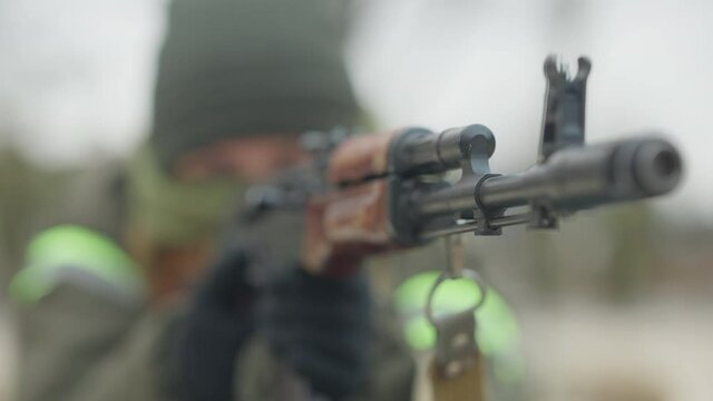 Close-up of gunpoint with blurred Caucasian soldier aiming at background. Close-up of army weapon with masked military woman training on polygon outdoors.